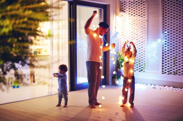 Ein Mann steht mit seinen beiden Kindern auf der Terrasse und spielt mit einer Lichterkette