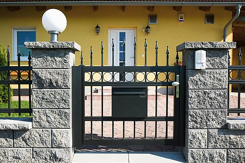 Gartentüre konvex in anthrazit mit Dekorringen und Lanze, mit passendem Briefkasten, Modell Venezia, vor gelbem Haus in Steinmauer integriert